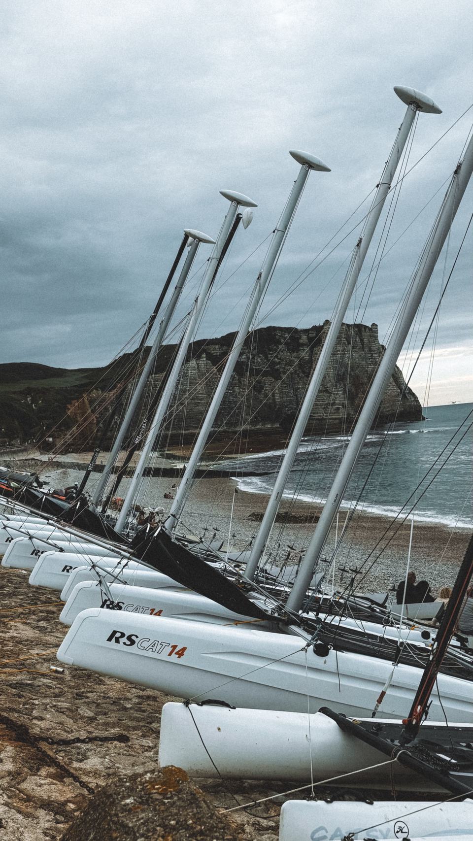 Catamarans de sport alignés sur une plage, falaise au fond